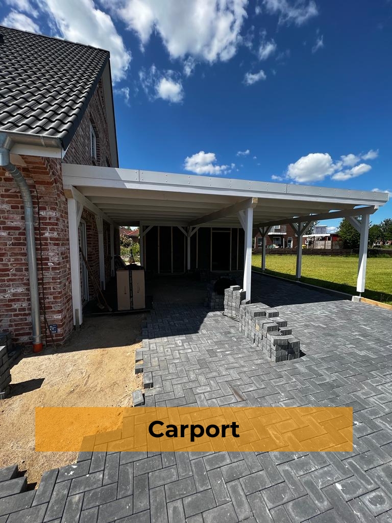 Ein freistehender, weiß gestrichener Holzcarport mit Flachdach, angebaut an ein Backsteinhaus, davor eine unfertige Pflasterung aus dunklen Steinen mit gelagerten Baumaterialien und einem orangefarbenen Banner mit dem Text „Carport“.