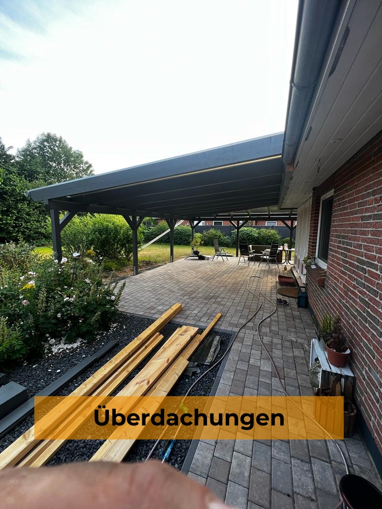 Große, dunkelgraue Terrassenüberdachung aus Holz an einem Klinkerhaus mit gepflasterter Terrasse und Gartenbereich, davor gelagertes Holz und im unteren Bereich ein orangefarbenes Banner mit dem Text „Überdachungen“.
