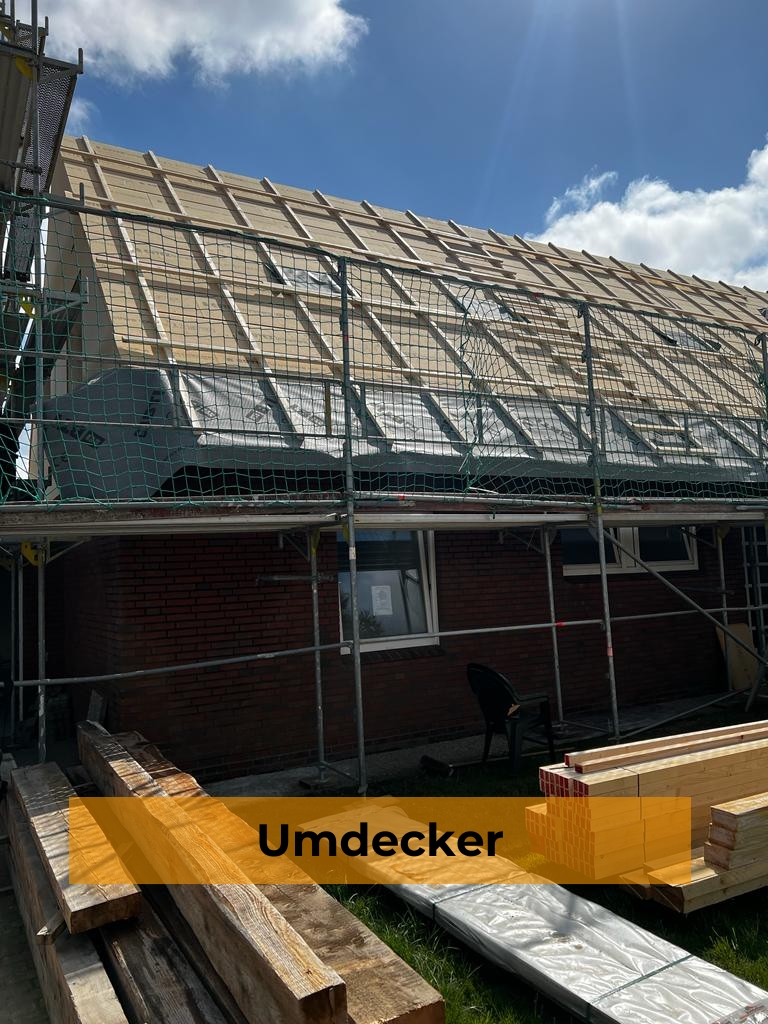 Dachstuhl-Sanierung an einem Klinkerhaus mit neu montierter Holzlattung hinter einem Baugerüst mit Sicherheitsnetz, davor gelagertes Bauholz und ein orangefarbenes Banner mit dem Text „Umdecker“.