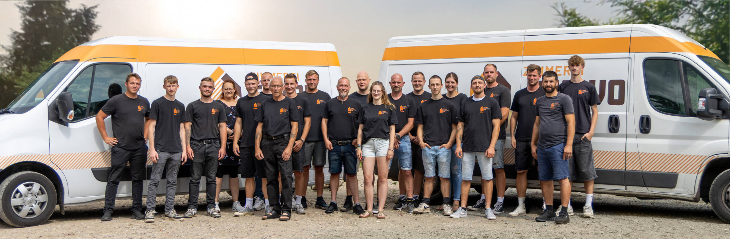 Gruppenfoto des Teams der Zimmerei SEKAVO in einheitlicher schwarzer Arbeitskleidung vor zwei weißen Firmen-Transportern im Freien.
